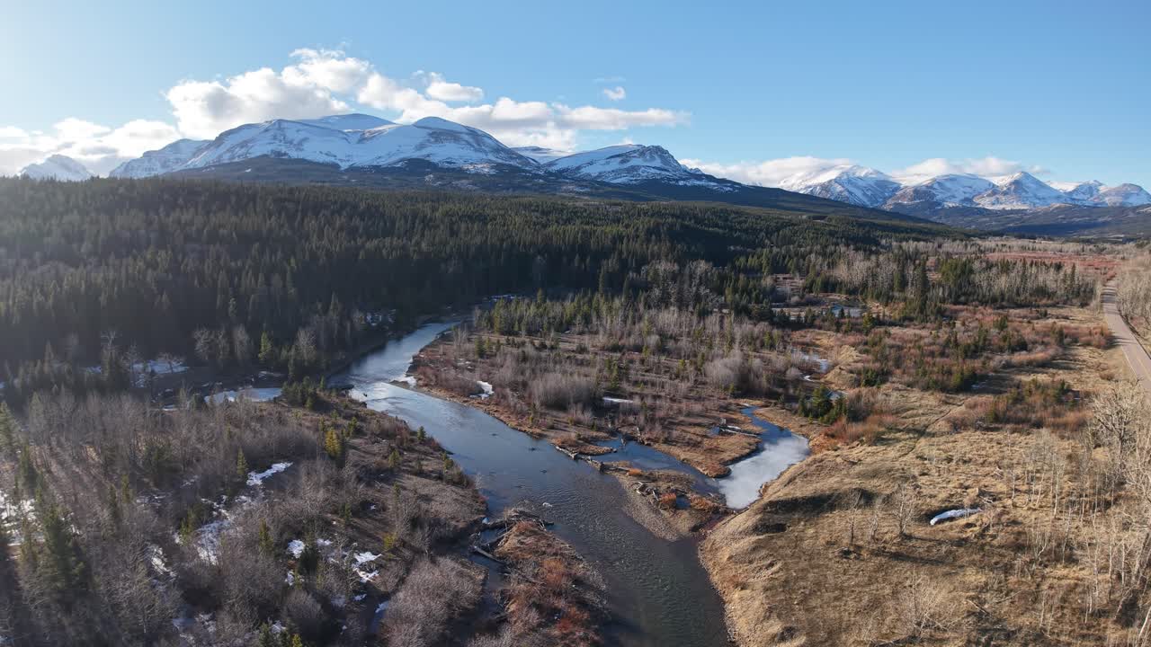 Ripresa con drone che segue un fiume tortuoso attraverso un'ampia valle circondata da foreste sempreverdi e prati bruni, con montagne innevate all'orizzonte vicino al Glacier National Park in Montana.