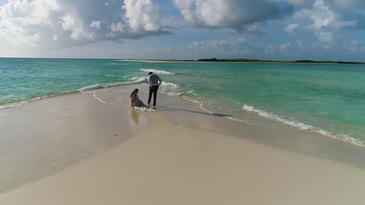 una pareja de recién casados se sienta en la playa de arena blanca para ver la puesta de sol en el mar, gira aérea alrededor de la isla caribeña