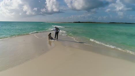 NEWLY-MARRIED-COUPLE-SIT-ON-WHITE-SAND-BEACH-WATCH-SEA-SUNSET,-AERIAL-TURN-AROUND-CARIBBEAN-ISLAND