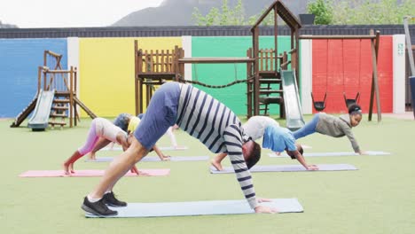 diverse male teacher and happy schoolchildren exercising on mats at school playground