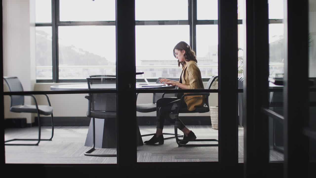 Young white businesswoman working alone in an office, seen through glass wall, full length, zoom out