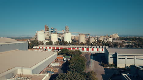 Aerial-shot-of-a-huge-cement-factory,-an-innovative-industrial-area-with-lots-of-hangars-and-trucks,-a-beautiful-area-with-trees,-push-in-shot-1