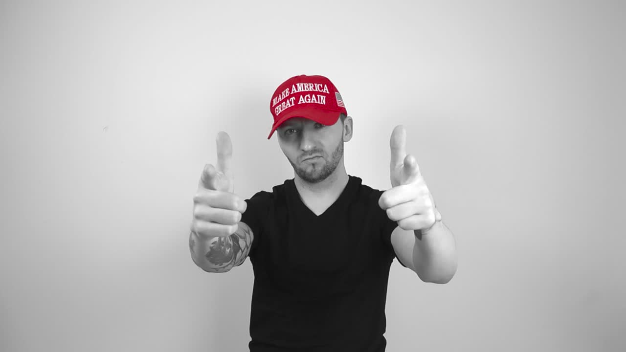 Grayscale footage of a tattooed man with a red MAGA Make America Great Again Trump hat lifting up his arms and points forward to the camera while nodding with a smile, showing he is happy for you.