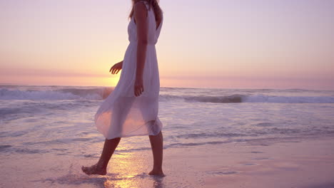 beautiful woman lifting white dress walking along shore line on  beach at sunset in slow motion red dragon