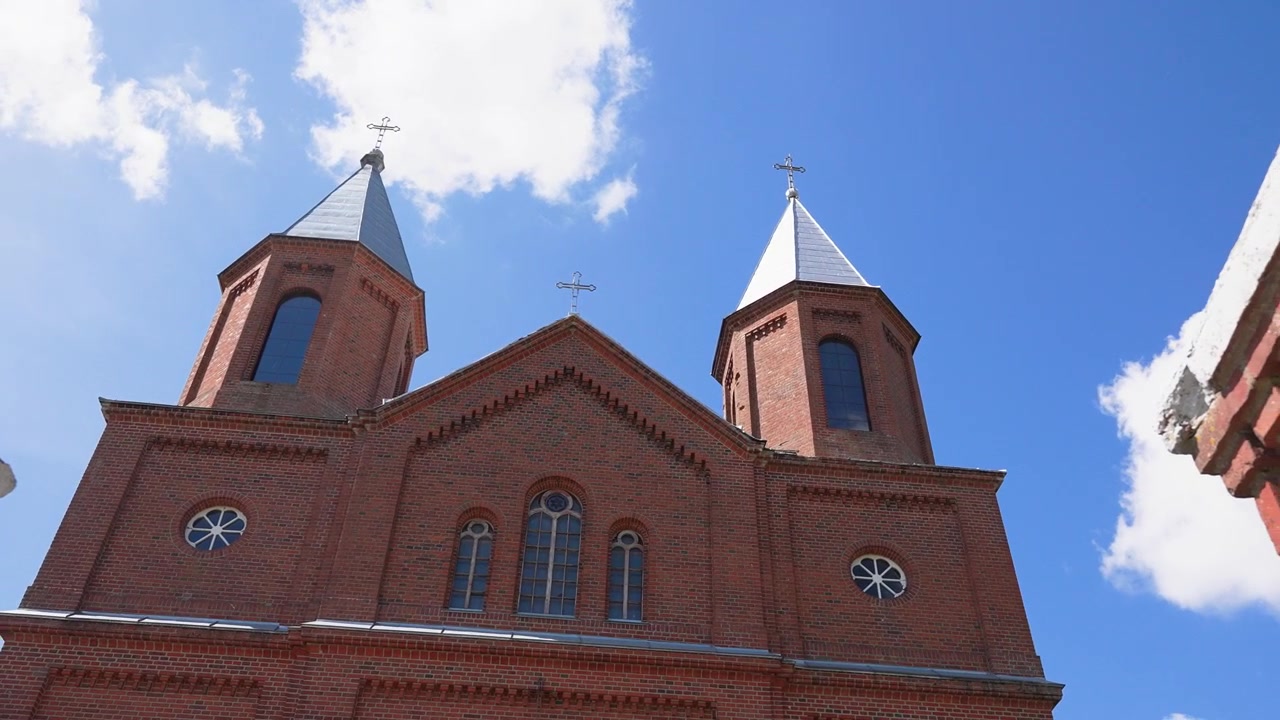 Imagens impressionantes em ângulo baixo com panorâmica para cima, revelando torres de igreja de tijolos vermelhos sob um céu azul vívido com nuvens dispersas. A luz do sol realça os detalhes arquitetônicos nesta imagem estável de grande angular.