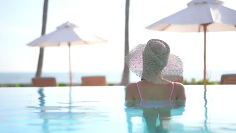 Espalda-De-Una-Mujer-Hermosa-Con-Sombrero-De-Sol-Parada-Dentro-Del-Agua-De-La-Piscina-En-Un-Salón-De-Resort-De-Isla-Tropical-Con-Sombrillas-De-Playa-Y-Tumbonas-En-El-Fondo,-Mano-A-Cámara-Lenta,-Plantilla-De-Vacaciones