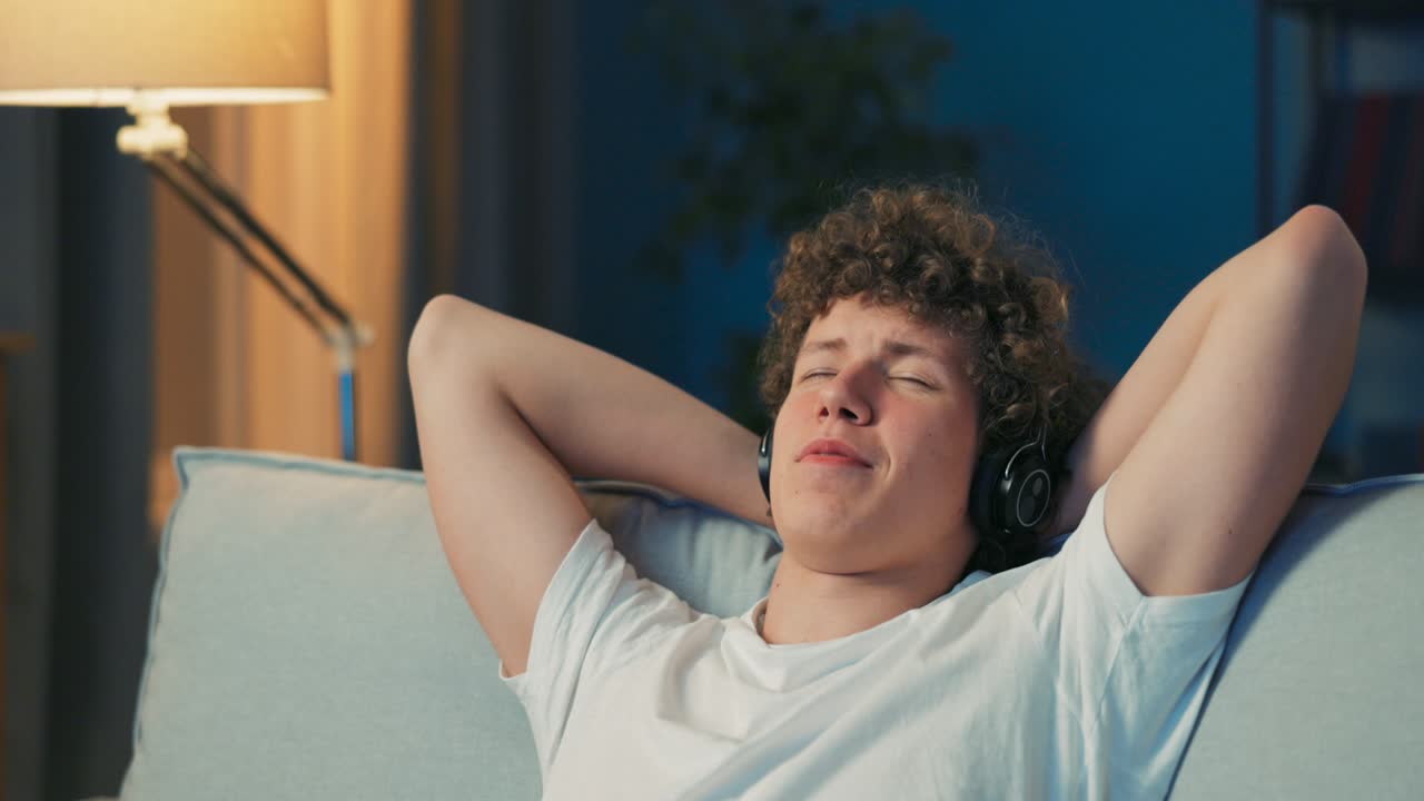 A men is resting on a sofa, hands behind head, listening to music on wireless headphones on ears, eyes closed, listening to a song, immersed in a dream, nodding, smiling
