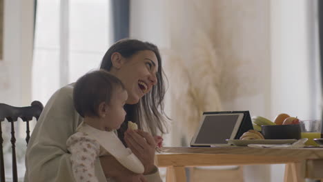 feliz hermosa mujer sentada en la mesa de la cocina y alimentando a su hija con plátano