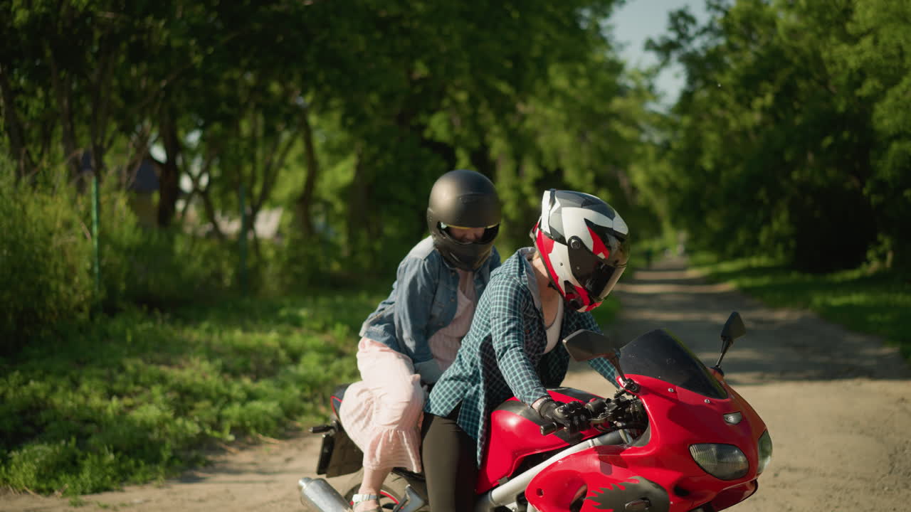 색 셔츠와 헬을 입은 한 여성이 은 파워 자전거에 앉아 있으며, 그녀의 친구는 분홍색 드레스와 파란색 재을 입고 조용하고 나무로 둘러싸인 포장 된 도로에 앉습니다.