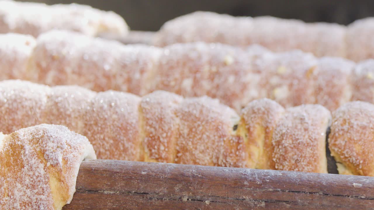 Sugary spiral pastries rotate on a wooden rod, cooked over an open grill in bright natural daylight. Close-up, shallow depth of field, steady camera
