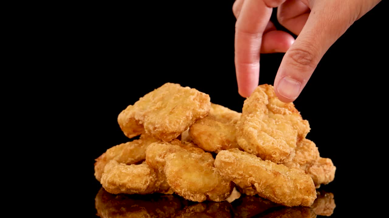 A hand reaches in to pick up a golden, crumbed chicken nugget from a neatly stacked pile on a reflective black surface, shot in slow motion