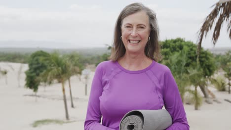 Portrait-of-happy-senior-caucasian-woman-holding-yoga-mat-and-smiling-on-beach,-in-slow-motion