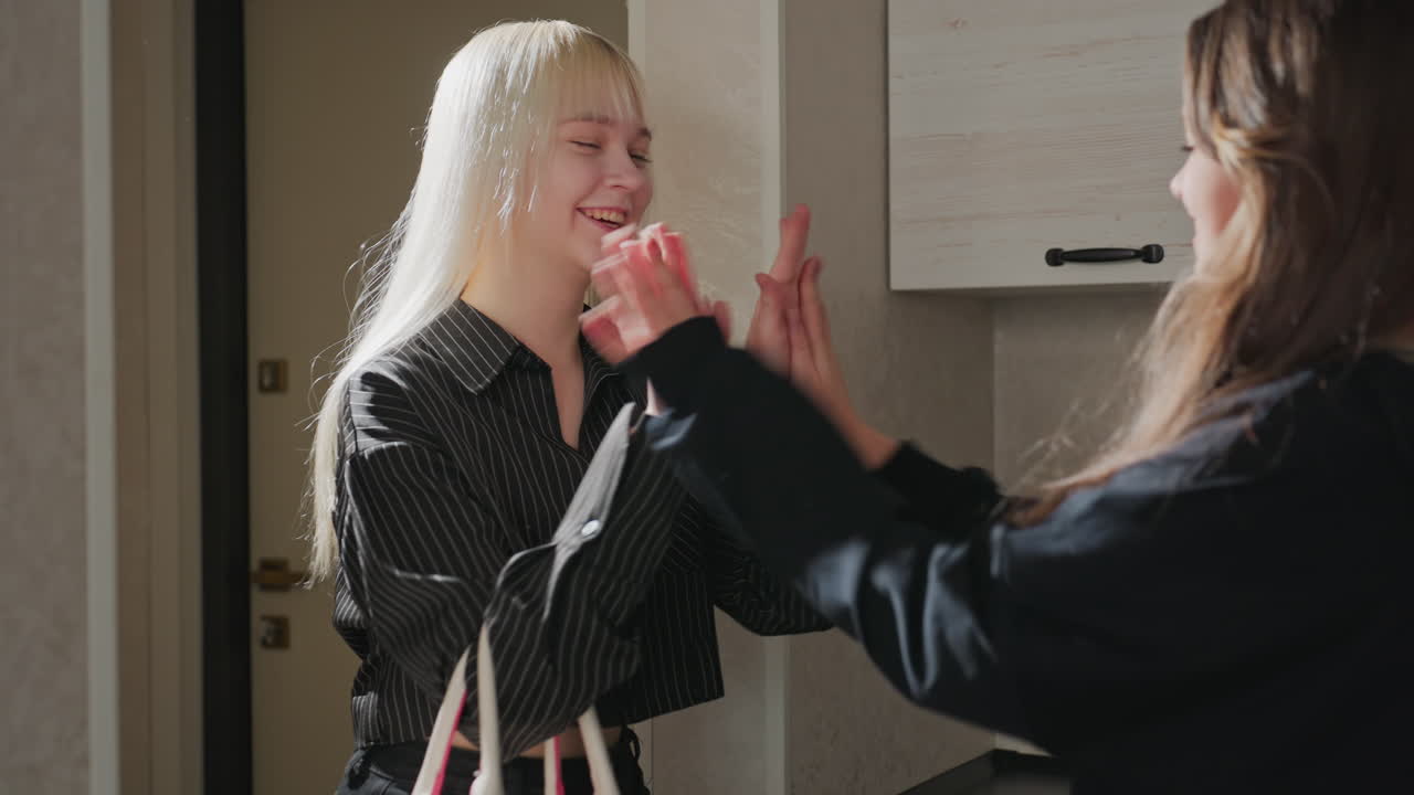 Close up of sisters laughing as they clap hands together near kitchen cabinets, light reflecting off blonde hair, capturing playful joyful sibling bonding and candid interaction