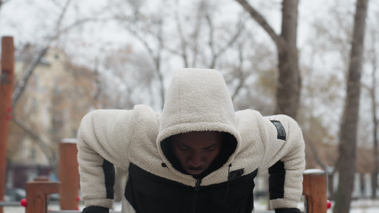 따뜻한 후디를 입은 젊은이가 눈이 내린 야외 환경에서 푸시업 운동을 하는 동안 초점을 맞추고 있으며, 그의 후드는 올려져 있으며, 그의 자세는 추운 날씨에 훈련하는 동안 집중력과 결단력을 반영합니다.