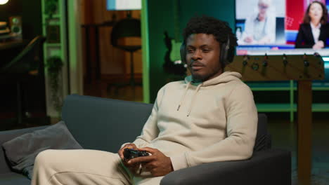 african american adult using headphones mic to talk with teammates while playing video games in cozy apartment. gamer chatting with internet friends while enjoying game on gaming console. camera a.