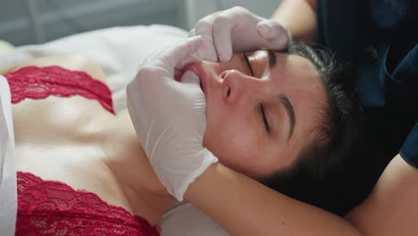 female participant on red top closes eyes gently as skin care expert wearing protective gloves dips fingernail inside mouth while other hand massages cheeks in clinical setting for gentle care
