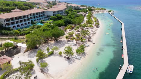 ascenso aéreo inclinado hacia abajo sobre la playa de arena blanca tropical con muelle y agua clara, curaçao