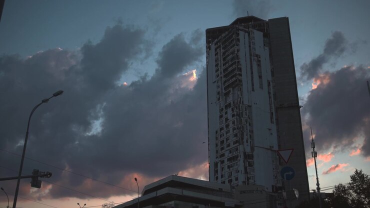 Uma torre de apartamentos alta e envelhecida domina a cena urbana ao lado de um semáforo iluminado, com primeiro plano escuro e nuvens densas ao anoitecer, apresentando uma suave luz de contorno laranja-rosada no horizonte.