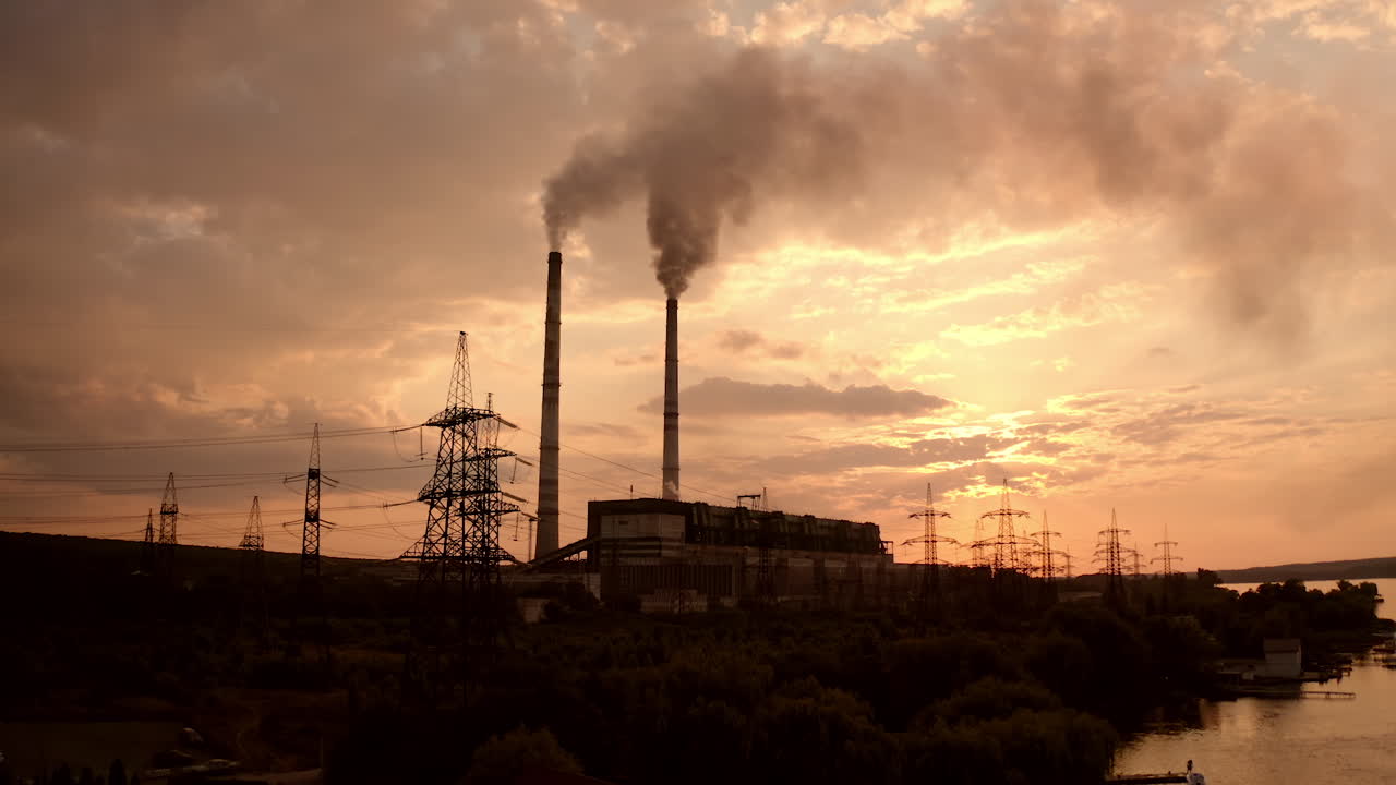 Dangerous industry pollutes the atmosphere. Harmful factory with large pipes smoking heavily near the river in the evening. Ecological disaster.