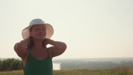 woman holds white hat preparing to wear it with confident gesture while standing in grassy hilltop overlooking winding river valley under soft morning light
