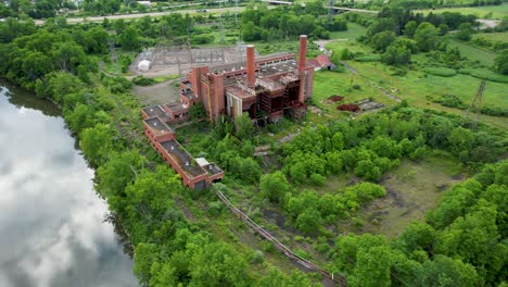 Abandoned-shut-down-Electric-Power-plant-along-a-river-in-green-overgrown-forest--aerial-turning-pan