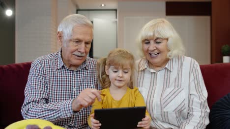 child girl shows something in laptop to grandparents, seniors couple learning how to use tablet