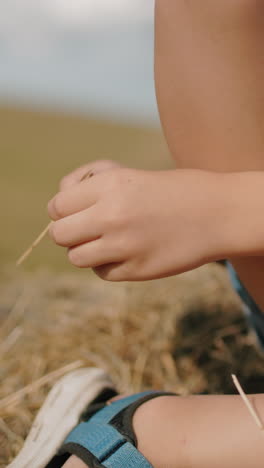 primer plano de las piernas del niño descansando en la bala de heno en el campo, usando sandalias casuales, el niño sostiene un pequeño pedazo de hierba mientras disfruta de un momento pacífico al aire libre en un clima cálido de verano bajo un cielo azul