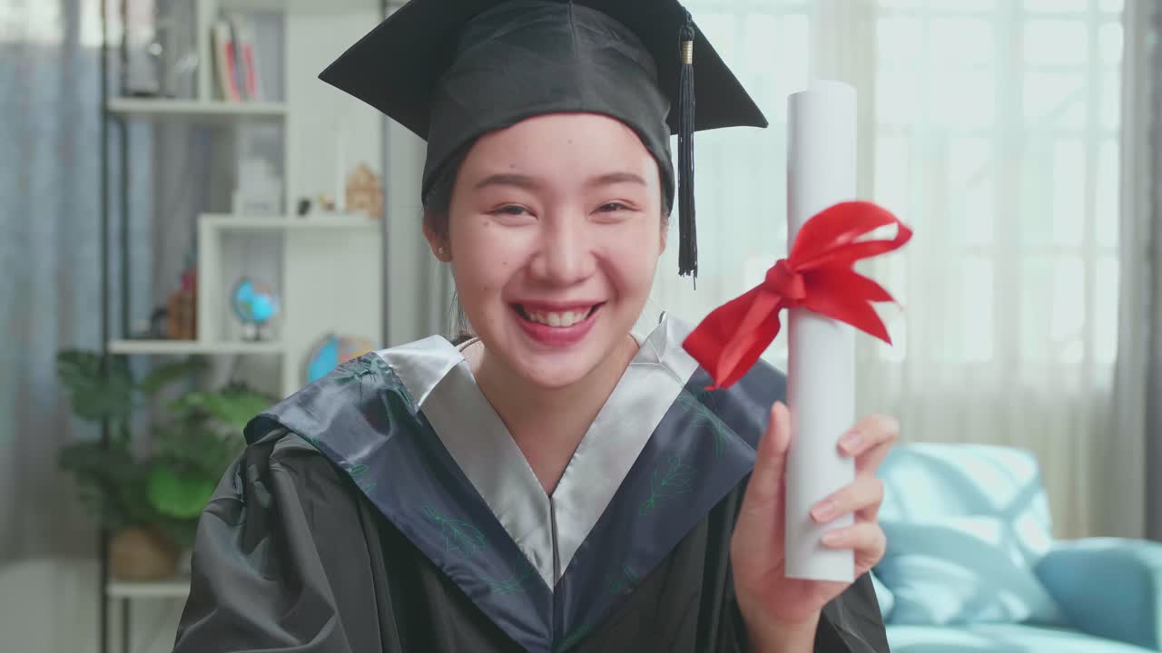 Excited Asian Woman Showing Off A University Certificate During An Online Video Call. Pretty Female Graduate Wearing A Graduation Gown And Cap Sitting On The Living Room