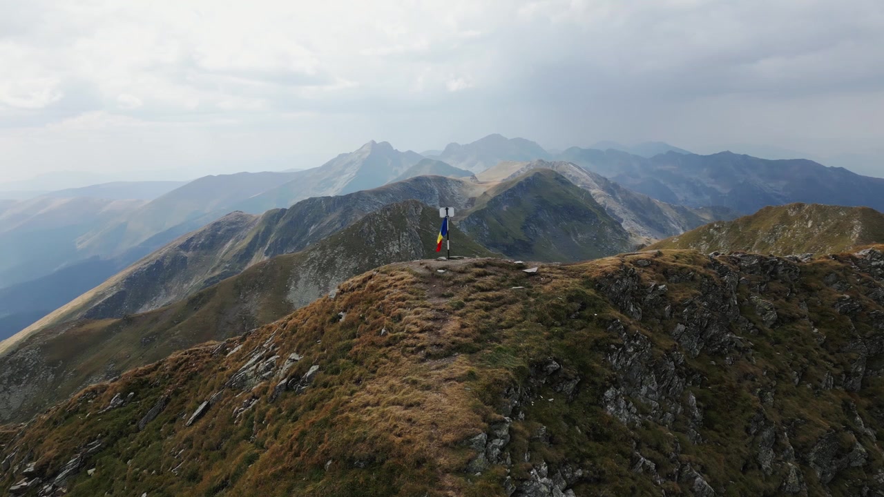 Orbita di un drone attorno a una grande cima di montagna con una bandiera rumena che sventola al vento in cima, Carpazi, Romania, Europa. Drone, estate.