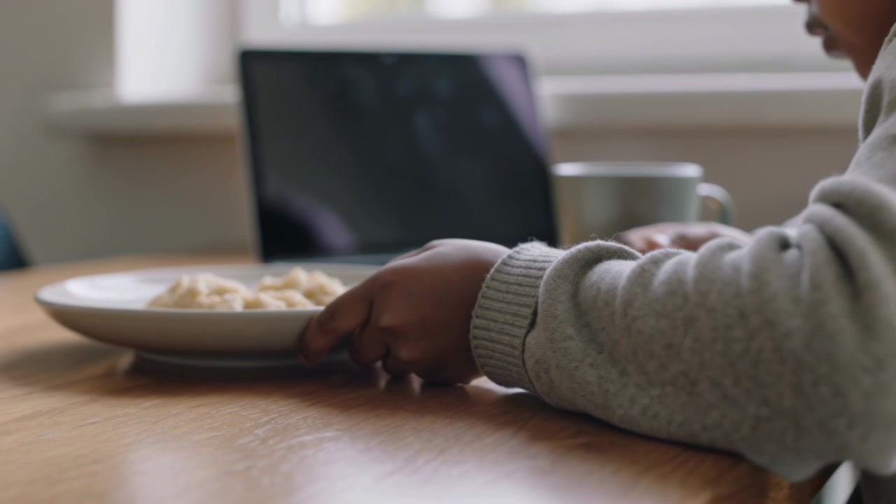 Leaning child grey sweater sliding plate closer to eat at home table with laptop, copy space