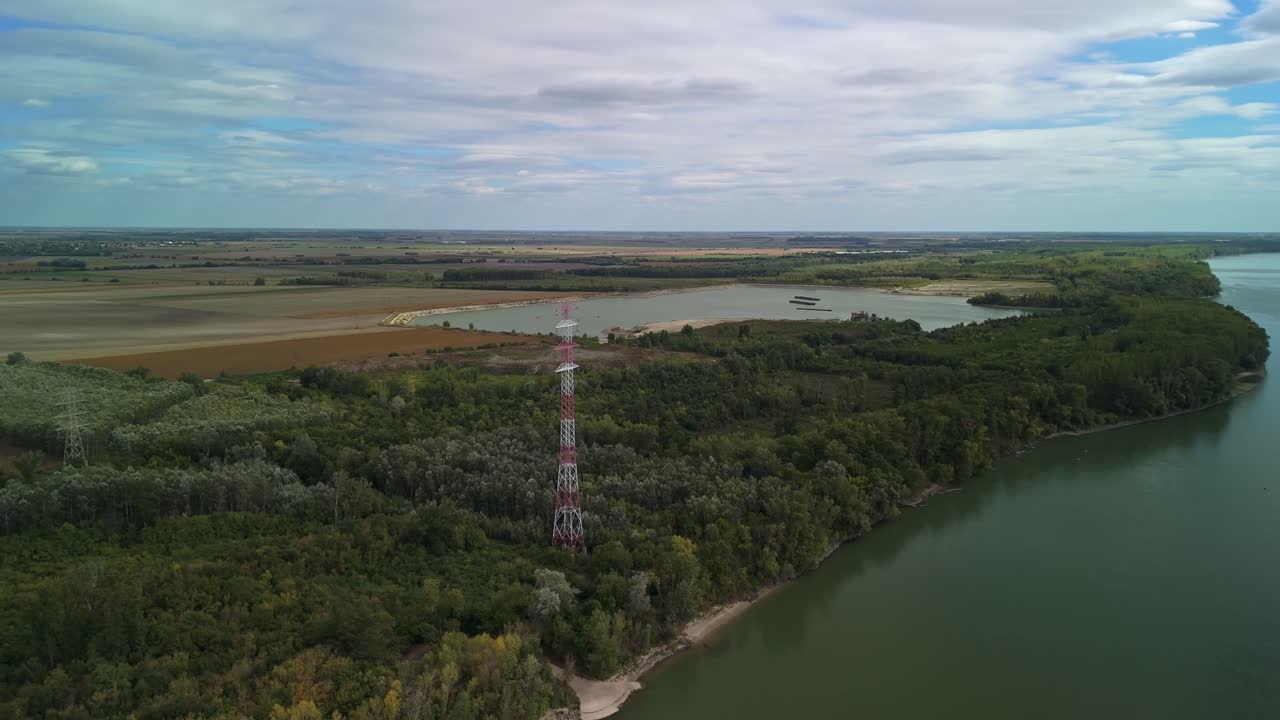 Flying towards a high electric pole surrounded by a mining lake, forests and agricultural lands along the Danube River near Szalkszentmárton in Hungary