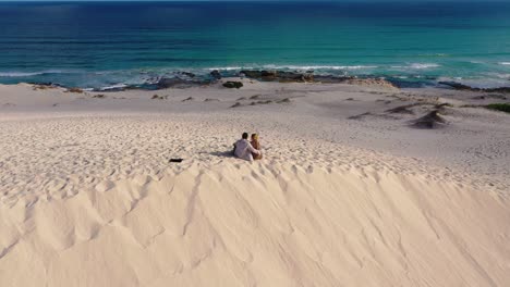 mixed couple sitting on a dune at a big sandy beach overlooking the ocean and sky while cuddling and holding hands in south africa filmed with drone while zooming out