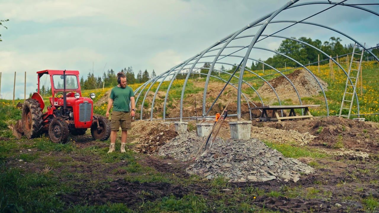 A Man Works Outdoors, Using A Tractor Equipped With A Shovel Attachment To Construct A Greenhouse Frame In Rural Area. Timelapse