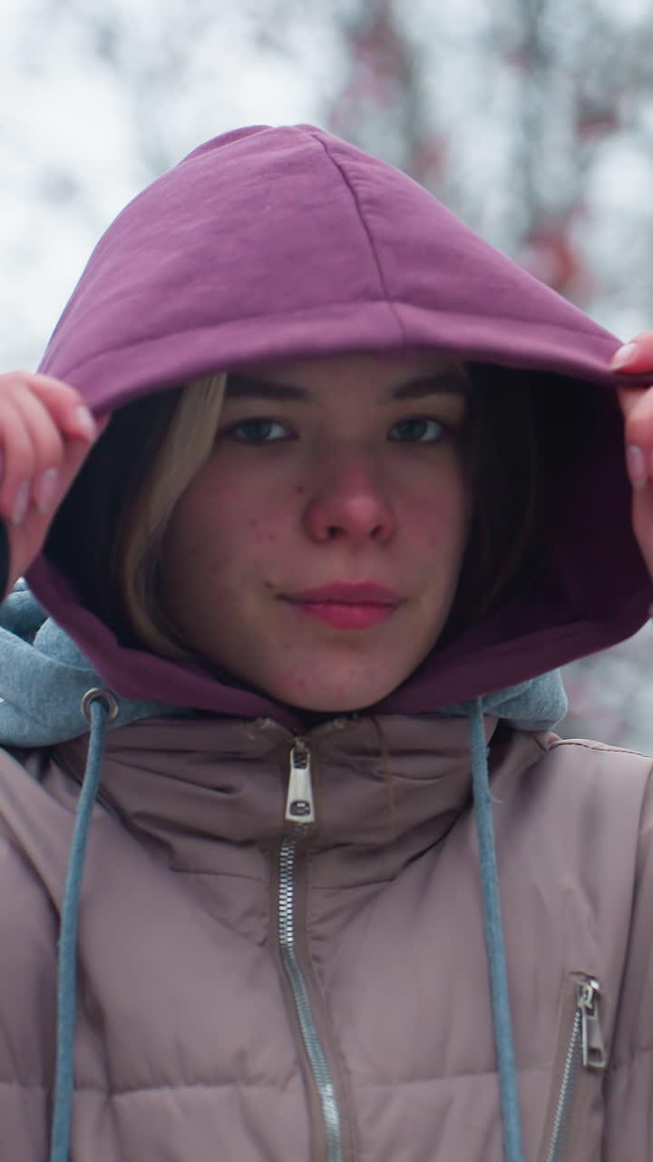 겨울 재을 입고 밖에서 자신감 있게 서있는 젊은 여자의 가까운 사진, 카메라에 대담한 시선으로 후디 모자를 조정, 나무와 가을 잎으로 흐릿한 배경