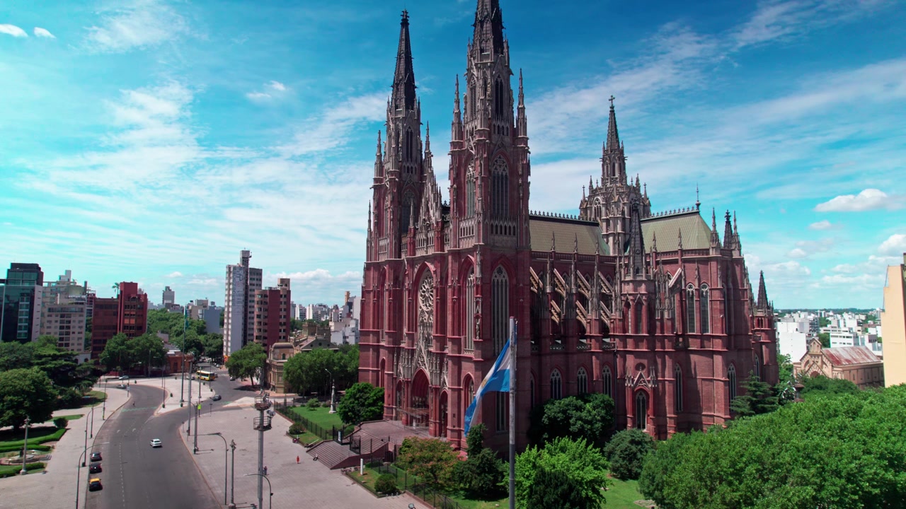 Ripresa aerea statica con drone che mostra il profilo laterale e le guglie della Cattedrale neogotica di La Plata, con la bandiera argentina che sventola in primo piano e il traffico cittadino a La Plata, Argentina