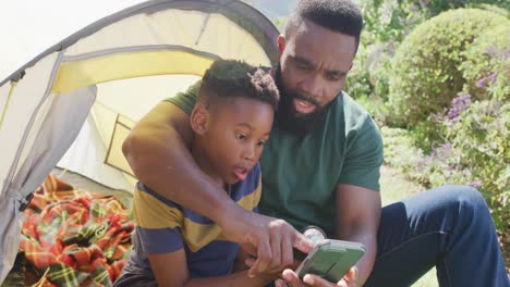 Glücklicher-Afroamerikanischer-Vater-Und-Sohn-Sitzen-Im-Zelt-Und-Benutzen-Das-Smartphone,-In-Zeitlupe