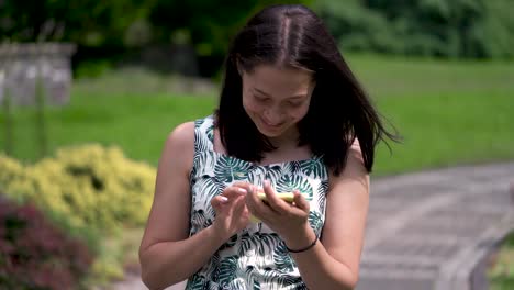 smiling asian girl in the summer dress walks along the track in the park. chatting in social networks. beautiful smile.