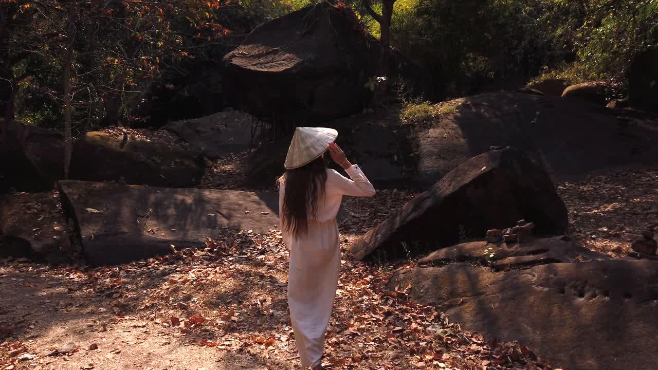 Young european woman traveler in long white dress and vietnamese hat walking in forest near huge boulders, big stones in Vat Phou – ruined Khmer Hindu Temple. Dry leaves, green trees around. Slow motion. Champassak, Laos, Asia.