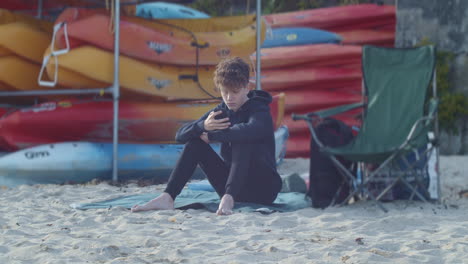 Adolescente-Se-Sienta-En-La-Playa-Con-Teléfono-Cerca-De-Alquiler-De-Kayak