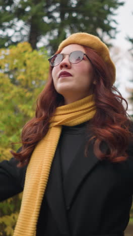 hermosa dama con el cabello rizado rojo se gira alegremente, colocando la mano en la cabeza, con boina amarilla, gafas de sol y silenciador en un jardín vibrante con colores de otoño