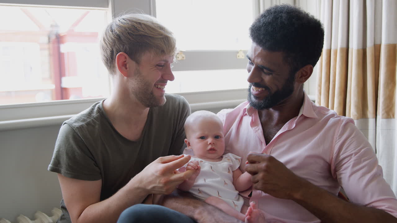 Portrait Of Loving Male Same Sex Couple Cuddling Baby Daughter On Sofa At Home Together