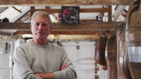 Portrait-Of-Senior-Male-Boxing-Coach-In-Gym-Standing-By-Old-Fashioned-Leather-Punching-Bags