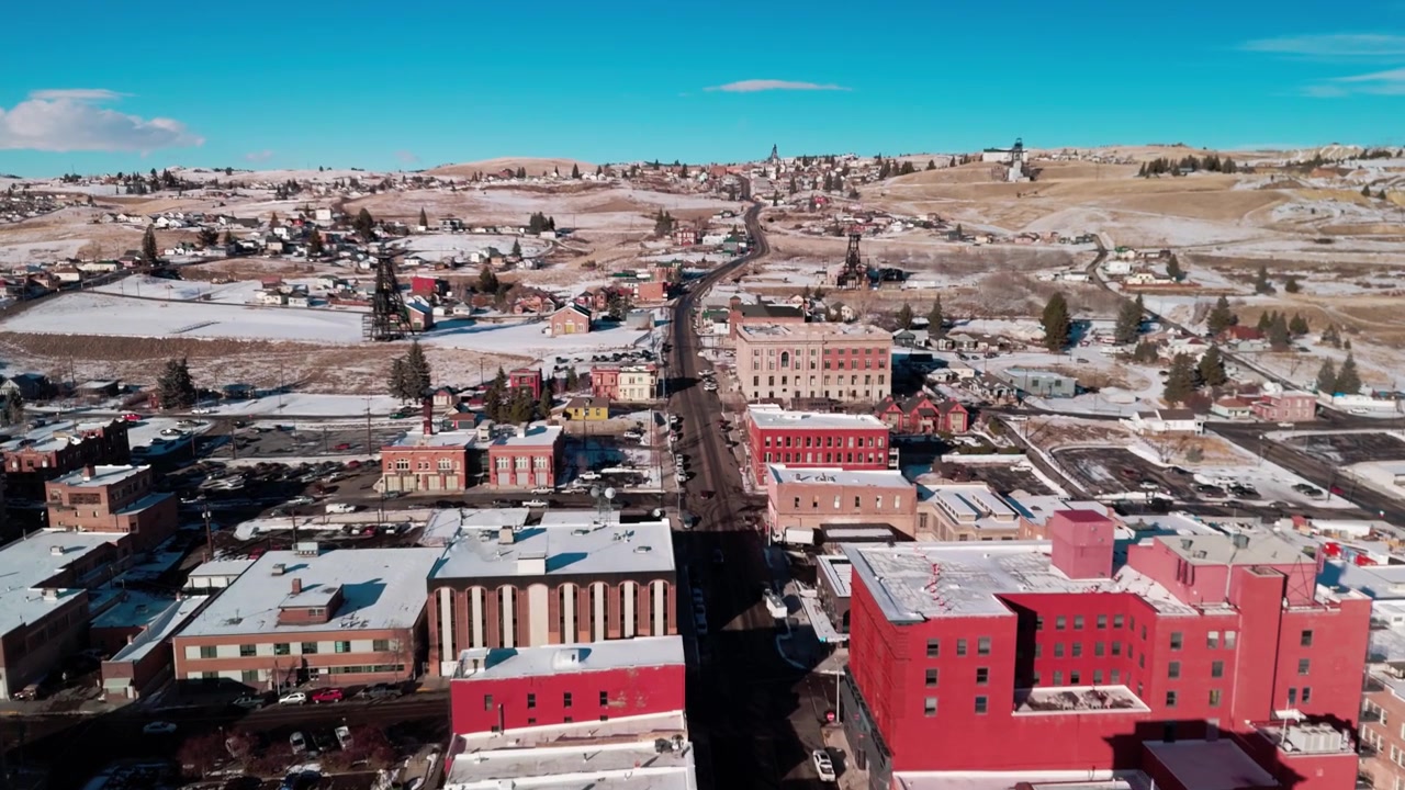 Vinterlig flyover fra luften over Butte, Montana, som illustrerer historiske industrielle gruvetårn og snødekte byblokker under en overskyet nordlig himmel fylt med vinterkulde.