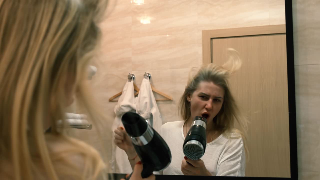 Giovane donna balla e canta con un asciugacapelli acceso davanti a uno specchio in piedi nel bagno. I capelli svolazzano al vento. 4k, slow motion