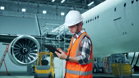 vista lateral de un ingeniero masculino asiático con casco de seguridad trabajando en una tableta mientras está de pie con aviones en el hangar