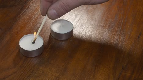 right hand uses burning wooden match to light two tea candles on table