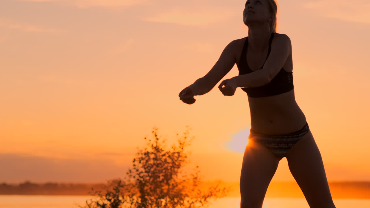 Middle distance volleyball girl in bikini waiting for the ball on the court at sunset gives forearm pass during a match on the beach in slow motion.