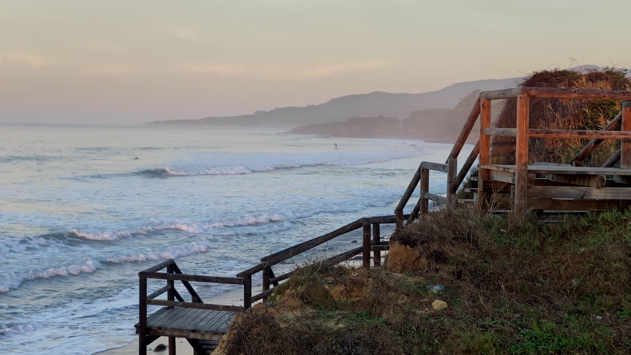A raised wooden platform stands above the sand at Hurricane, Tarifa, facing soft evening waves and a hazy pastel horizon