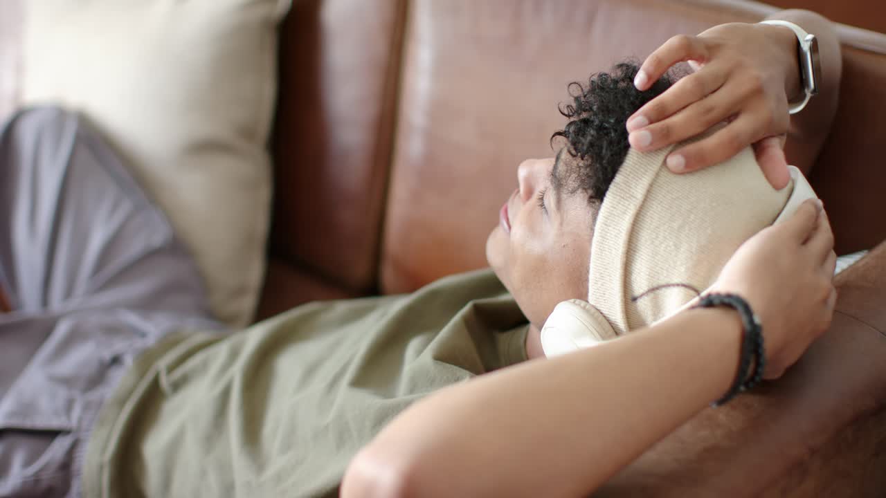 Un adolescente sdraiato sta sistemando un cappello beige con cuffie bianche su un divano marrone, assicurandosi che calzi bene.