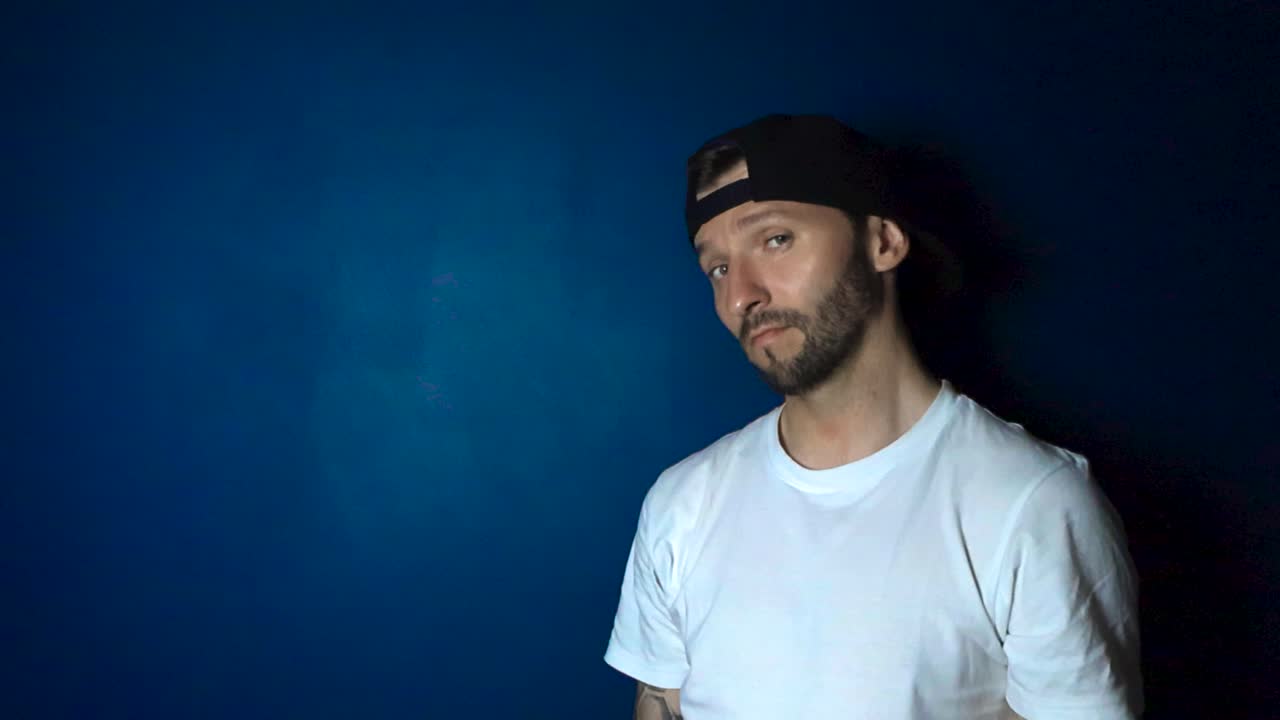 Tattooed man with a white shirt and a black reversed cap hat lifting up a softbox lighting gear in front of a blue wall and nodding with approval. Influencer showing a studio light that is proper